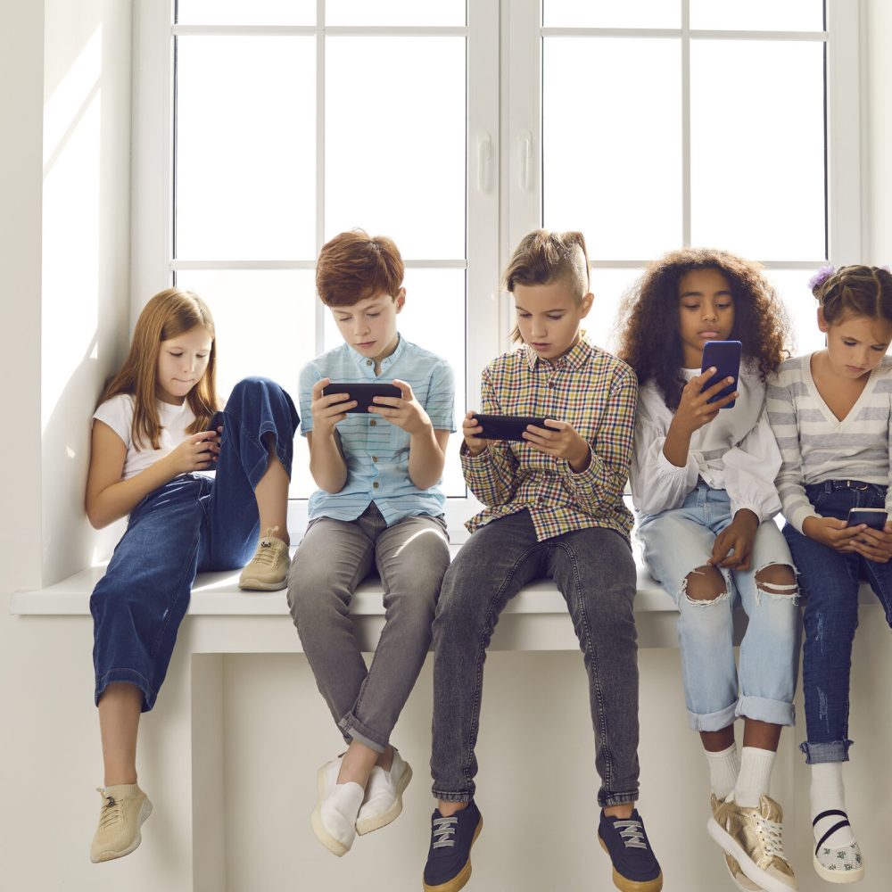 Group of children of different nationalities sit in a row on the windowsill and with lack of emotion play online games or read social networks on mobile phones. Technologies that spoil childhood.