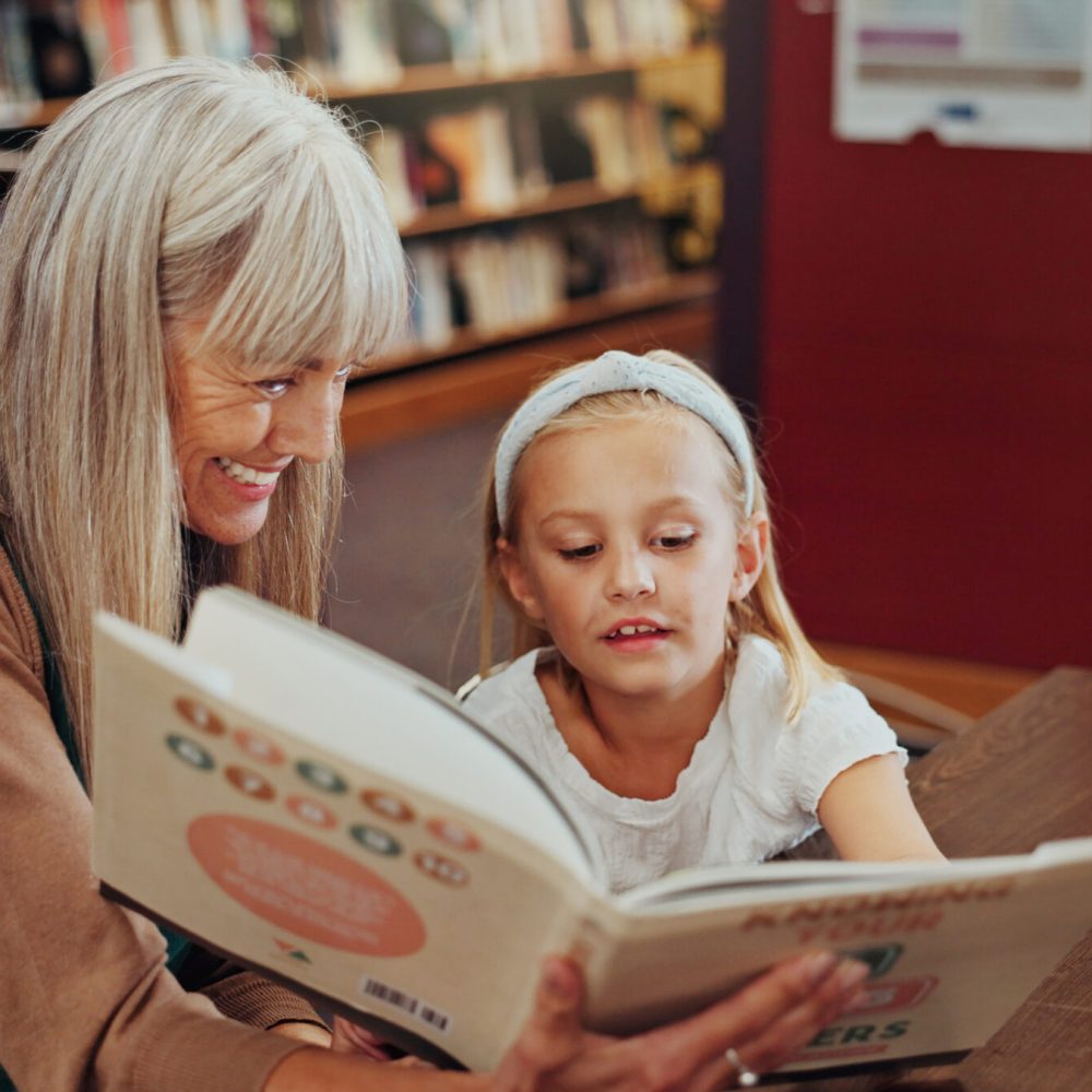 Grandmother, reading book and girl in library, storytelling and bonding together for child development. Mature woman, kid or support for education, literacy and hobby with family and english language